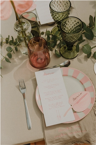 Detalle de puesta en mesa con menú de boda impreso, marcasitios en forma de hoja, vajilla con plato a rayas rosa, copas de cristal verde y rosa, y servilleta bordada.