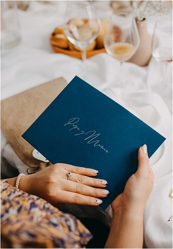Mujer sosteniendo un sobre azul marino con caligrafía blanca que dice “Papá y Mamá”, en una mesa decorada para una celebración.