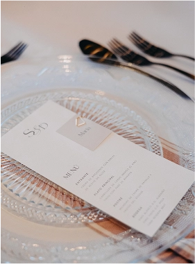 Minuta de boda con diseño elegante y tipografía clásica colocada sobre un plato de cristal tallado, con cubertería negra y servilleta nude de fondo. El diseño incluye las iniciales “S & D” y un gráfico geométrico en relieve con la palabra "Menu".