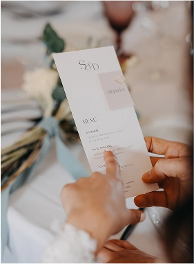 Persona sosteniendo una minuta de boda elegante con las iniciales "S & D", presentada en formato vertical sobre fondo blanco con detalles gráficos en tono nude. Al fondo, un ramo atado con cinta azul y una mesa decorada con cristalería y flores.