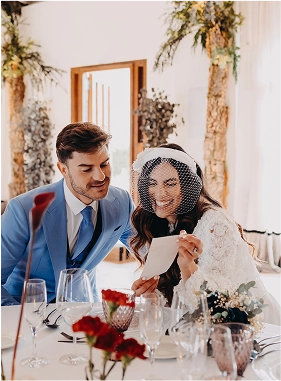 Pareja de recién casados sentados en la mesa del banquete revisando la papelería de la boda entre sonrisas. Ella lleva un vestido de encaje y tocado con redecilla, y él viste traje azul. La decoración incluye copas de colores, centros de flores rojas y vegetación.