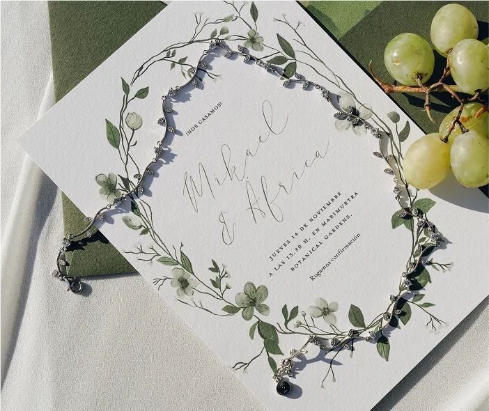 Invitación de boda personalizada con ilustraciones de ramas y flores verdes sobre papel texturizado, acompañada de un colgante plateado en forma de corona y un racimo de uvas verdes. La papelería está colocada sobre telas en tonos blanco y verde oliva.