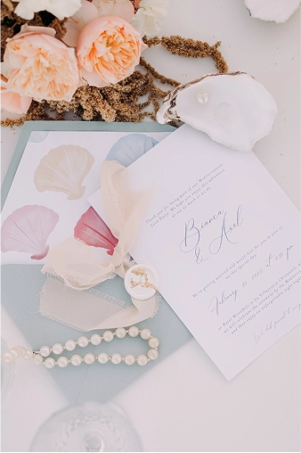 Invitación de boda con tipografía elegante en azul sobre papel blanco, acompañada de un sobre azul pastel con ilustraciones de conchas marinas en tonos rosa, crema y lavanda. La escena incluye una pulsera de perlas, lazos de tela y flores melocotón, sobre una mesa decorada con conchas y detalles orgánicos, evocando una temática costera y romántica.